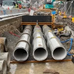 Four large concrete pipes are being installed underground at a construction site, with heavy machinery and safety barriers visible in the background.