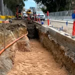 A construction site with an open trench, exposed orange pipe, construction vehicles, safety barriers, and warning flags along a partially completed road.