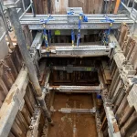Steel and wooden shoring supports reinforce an excavation site with wet soil at the bottom, surrounded by construction materials and safety railings.