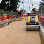 A construction worker operates a road roller on a sandy road under repair, with safety barriers and fencing on both sides.