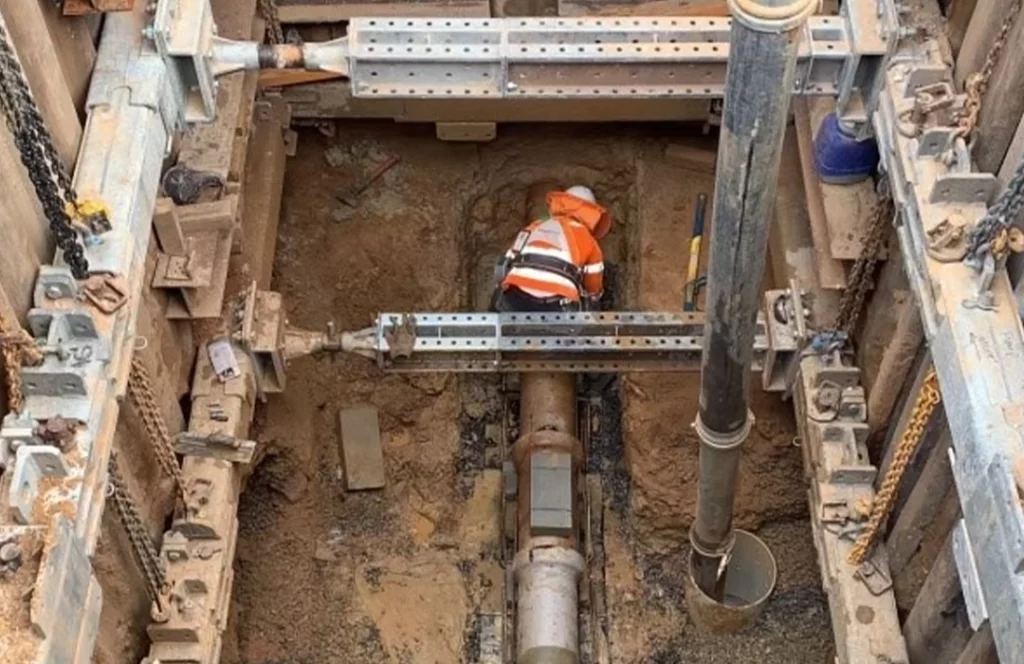 A construction worker in a fluorescent vest works on a pipeline installation inside a deep, reinforced excavation site filled with soil and metal supports.