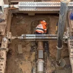 A construction worker in a fluorescent vest works on a pipeline installation inside a deep, reinforced excavation site filled with soil and metal supports.