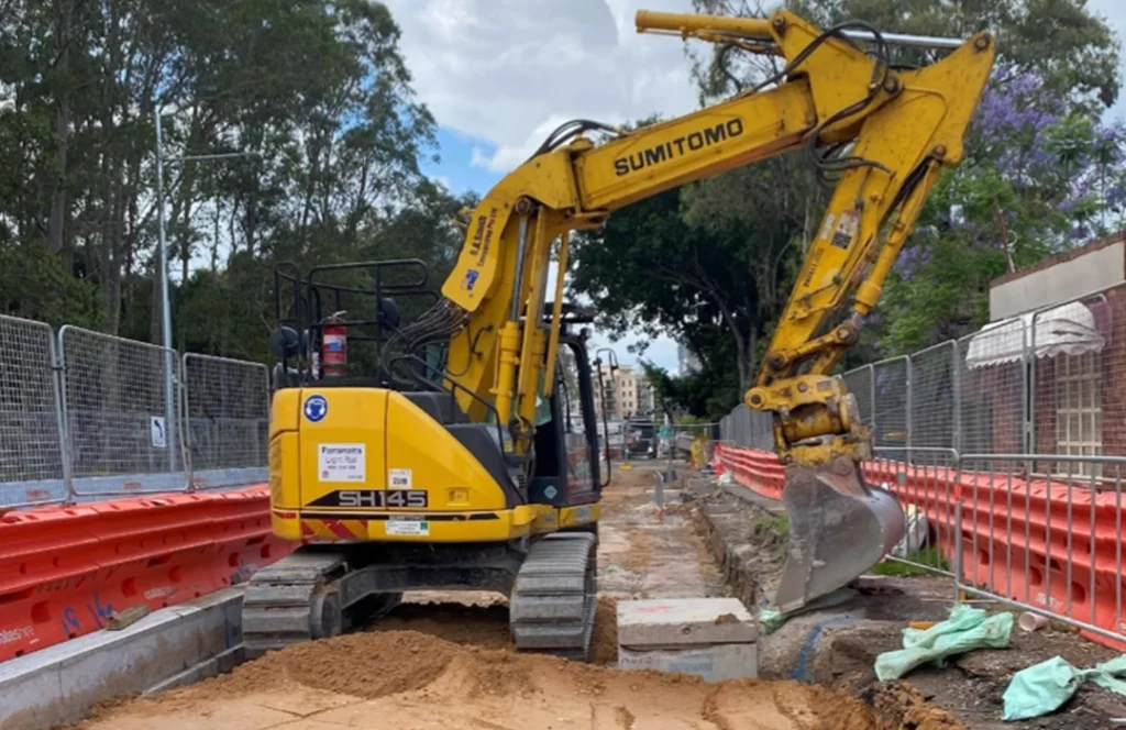 A yellow excavator is parked on a construction site, surrounded by orange barriers and fencing, with trees and buildings in the background.