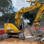 A yellow excavator is parked on a construction site, surrounded by orange barriers and fencing, with trees and buildings in the background.