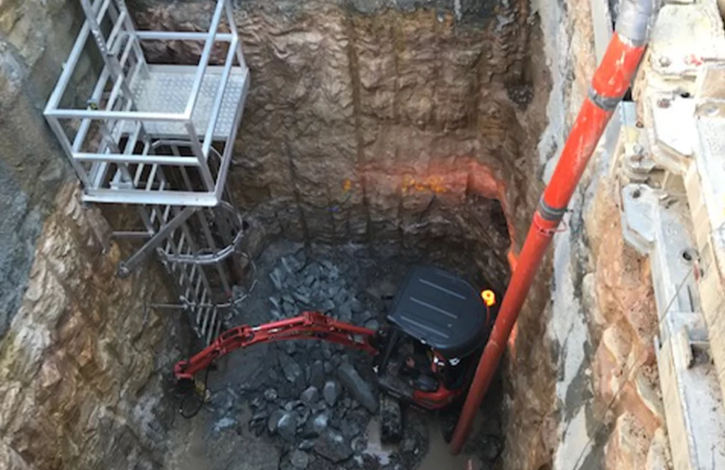 An excavator operates at the bottom of a deep construction pit with a metal access ladder and a large red pipe along the wall.