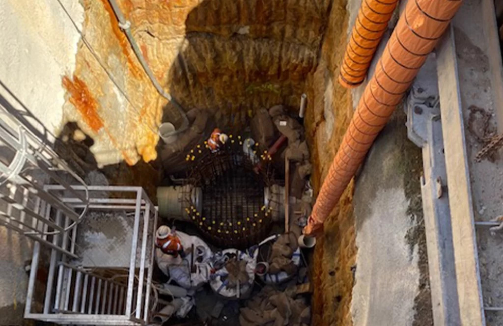Aerial view of workers in protective gear at the bottom of a deep construction pit, surrounded by scaffolding, pipes, and excavation equipment.