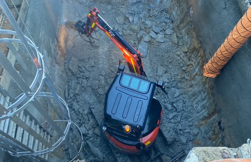 A small excavator with a red arm digs in a deep, narrow pit surrounded by concrete walls and loose rubble, near a metal ladder and an orange protective cover.