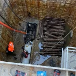 A construction worker wearing an orange safety vest stands in a deep rectangular pit with machinery, pipes, and equipment stacked inside.