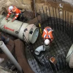 Three construction workers in safety gear install large concrete pipes and metal reinforcement in an excavated trench at a construction site.