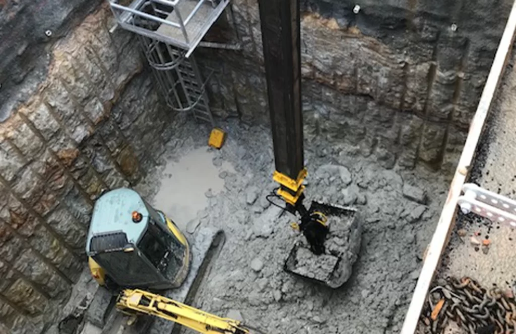 Excavator digging a deep, rectangular pit with rocky walls; metal stairs are installed in one corner for access.