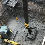 Excavator digging a deep, rectangular pit with rocky walls; metal stairs are installed in one corner for access.
