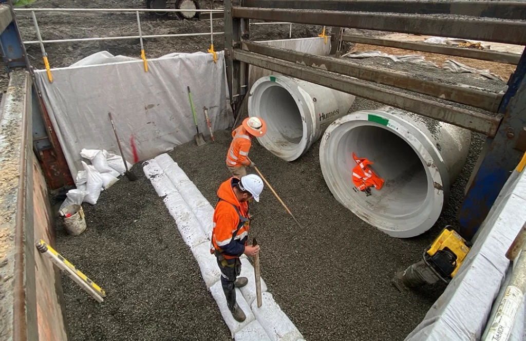 Three construction workers install large concrete pipes in a trench, surrounded by gravel, tools, safety barriers, and sacks.
