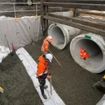 Three construction workers install large concrete pipes in a trench, surrounded by gravel, tools, safety barriers, and sacks.