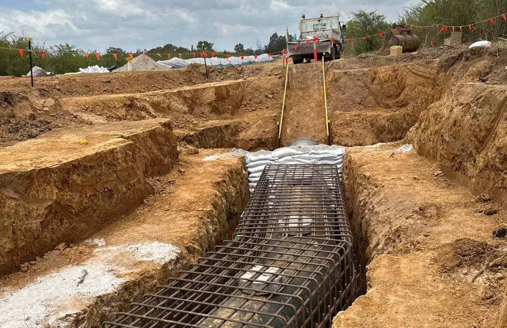 A large pipeline covered with steel rebar is being installed in a trench at a construction site with a truck and equipment in the background.