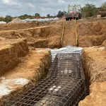 A large pipeline covered with steel rebar is being installed in a trench at a construction site with a truck and equipment in the background.