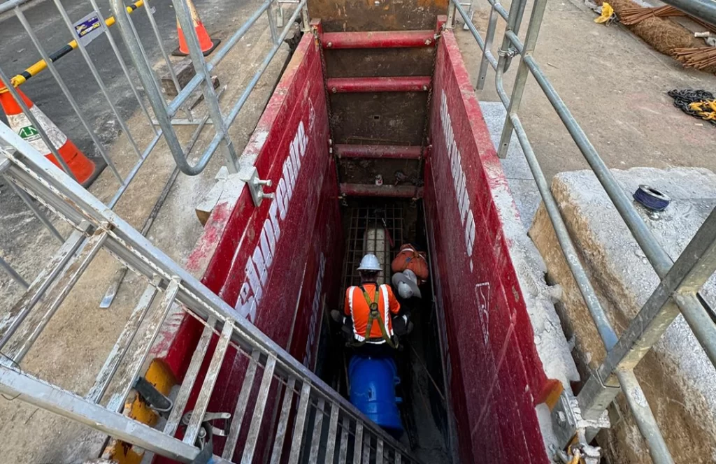 Two construction workers wearing safety gear are working inside a deep trench secured with red barriers and metal ladders at a construction site.