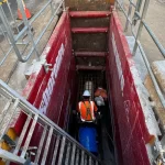 Two construction workers wearing safety gear are working inside a deep trench secured with red barriers and metal ladders at a construction site.