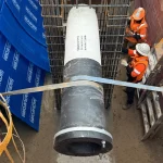 Two construction workers in safety gear stand in a trench beside a large concrete pipe being lowered into place with straps and scaffolding around.