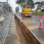 A deep trench runs along a road under construction, with safety barriers, orange cones, and a Komatsu excavator on site.