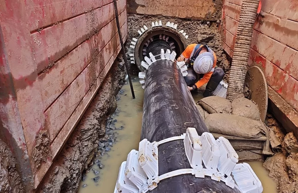 A worker in safety gear installs or inspects a large black pipeline in a muddy trench, surrounded by construction barriers and sandbags.