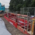 Construction site with red trench box, fencing, and an excavator in the background. Workers are present near the equipment, and trees are visible behind the site.