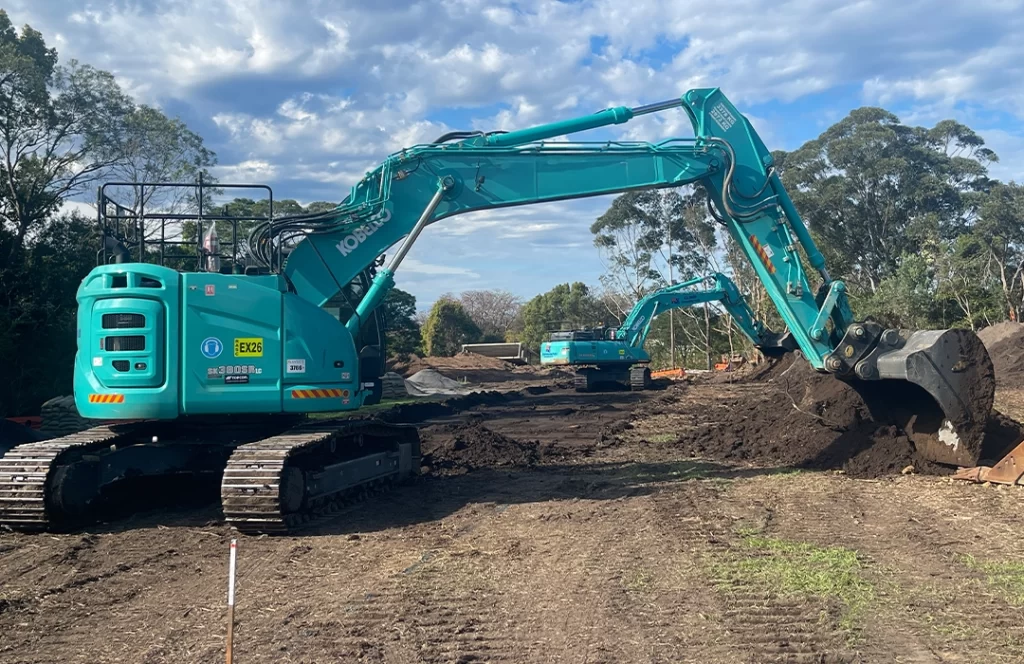 Two teal excavators work on a dirt construction site surrounded by trees and a partly cloudy sky.