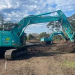 Two teal excavators work on a dirt construction site surrounded by trees and a partly cloudy sky.