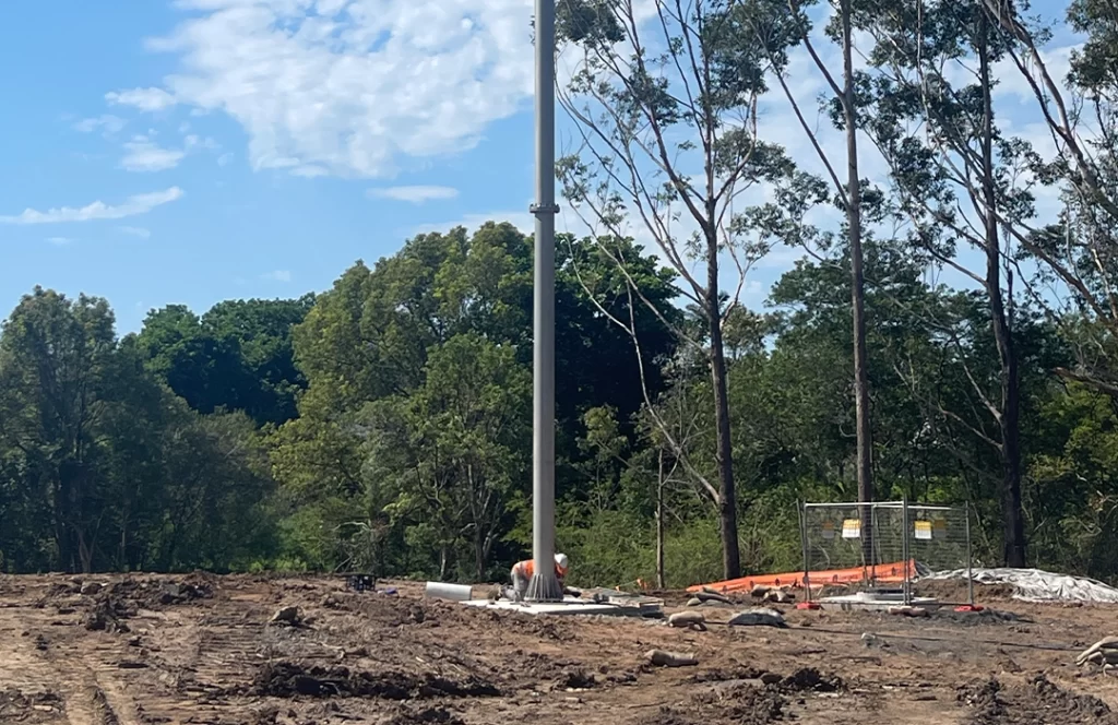 A worker in a safety vest and helmet stands near a pole on a cleared construction site bordered by trees under a partly cloudy sky.