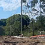 A worker in a safety vest and helmet stands near a pole on a cleared construction site bordered by trees under a partly cloudy sky.