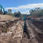 A construction site with deep trenches, excavators, and workers in safety gear; red safety flags line the trench edges, surrounded by trees under a partly cloudy sky.
