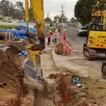 A construction site with workers and excavators digging a trench along a roadside, surrounded by equipment, pipes, and safety barriers.