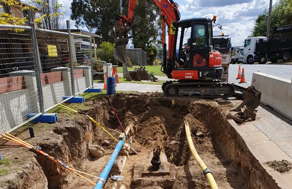A small excavator is digging a trench in a residential area, exposing pipes and cables, with safety barriers and traffic cones set up nearby.