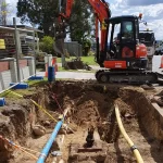 A small excavator is digging a trench in a residential area, exposing pipes and cables, with safety barriers and traffic cones set up nearby.