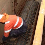 A construction worker in a high-visibility jacket and helmet installs steel reinforcement bars over a black membrane inside a trench on a worksite.