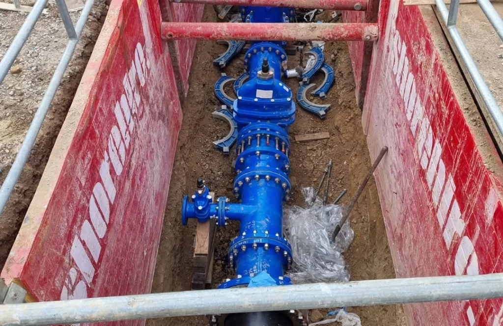 A newly installed blue water valve assembly is positioned in a trench with red support barriers on either side, surrounded by bolts and construction materials.