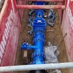 A newly installed blue water valve assembly is positioned in a trench with red support barriers on either side, surrounded by bolts and construction materials.
