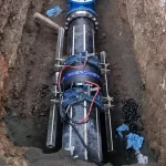A large black pipe with blue fittings and electronic sensors is installed in a trench, surrounded by soil and scattered debris.