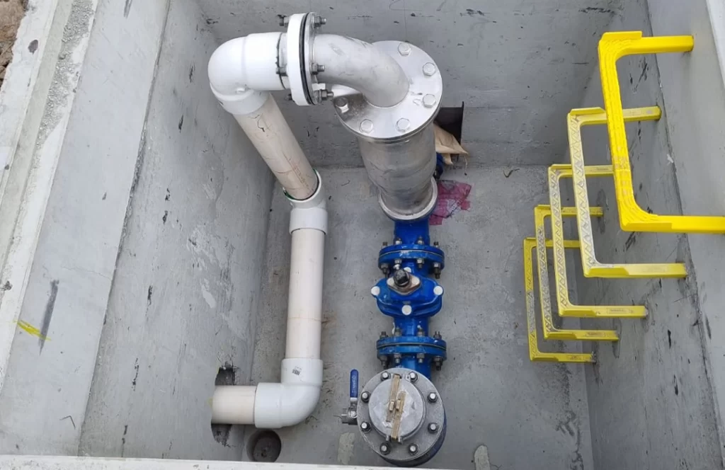 A concrete pit containing white PVC and blue metal pipes, valves, a cylindrical filter, and yellow metal ladder steps mounted on the wall.