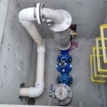 A concrete pit containing white PVC and blue metal pipes, valves, a cylindrical filter, and yellow metal ladder steps mounted on the wall.