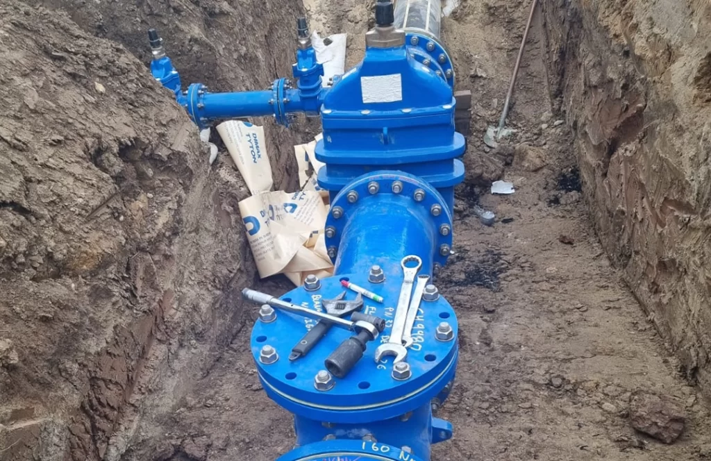 Large blue pipes and valves are installed in a trench, with tools and sandbags placed on top, indicating ongoing utility or water line construction work.