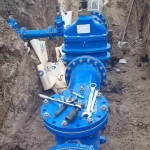 Large blue pipes and valves are installed in a trench, with tools and sandbags placed on top, indicating ongoing utility or water line construction work.