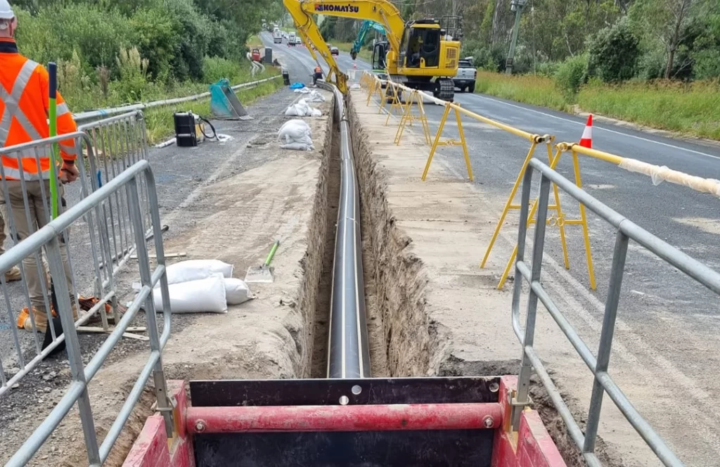A construction site with a long, narrow trench running alongside a road, containing a large pipe. An excavator and workers are present, with safety barriers set up.