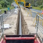 A construction site with a long, narrow trench running alongside a road, containing a large pipe. An excavator and workers are present, with safety barriers set up.
