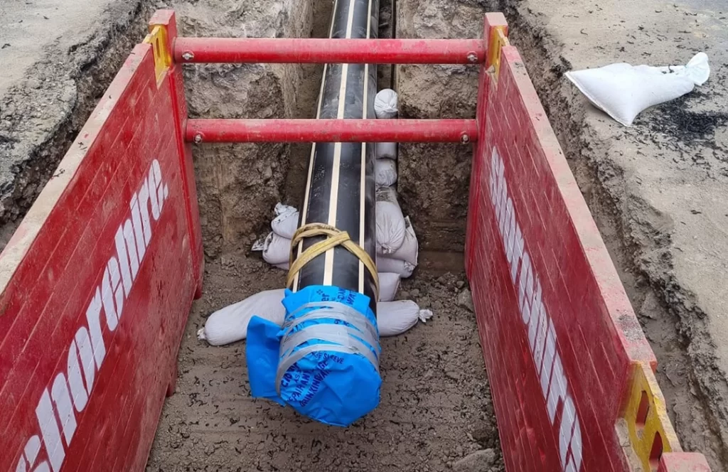 A large black pipe wrapped at one end with blue material is secured in a trench, supported by sandbags and red safety barriers on both sides.