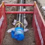 A large black pipe wrapped at one end with blue material is secured in a trench, supported by sandbags and red safety barriers on both sides.