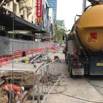 A construction worker digs in a street trench near a vacuum truck, with barriers and safety fencing along a city sidewalk.