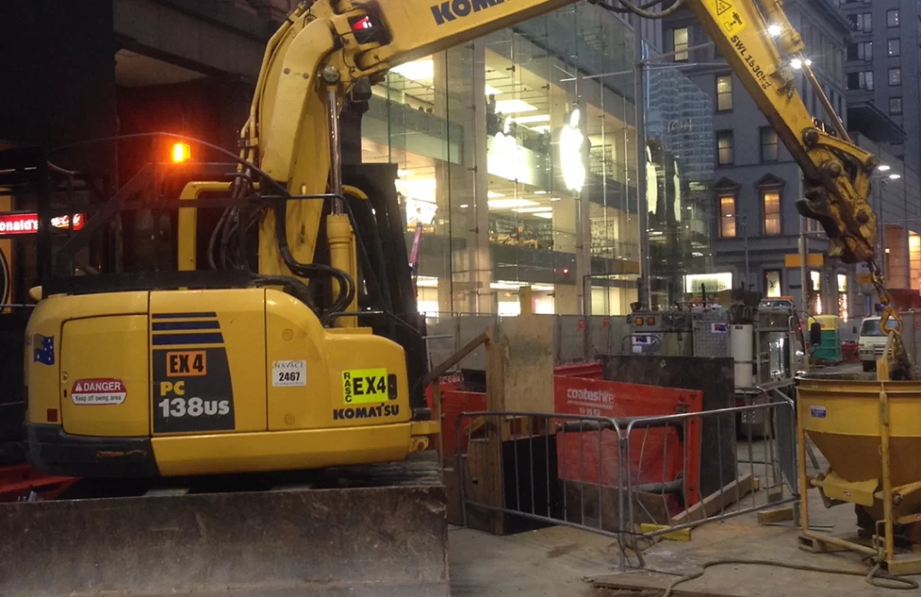 A yellow Komatsu excavator is parked at a construction site near a glass-fronted building with an Apple logo in an urban area.