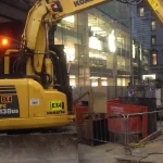 A yellow Komatsu excavator is parked at a construction site near a glass-fronted building with an Apple logo in an urban area.