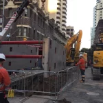 Construction workers in safety gear operate heavy machinery and install concrete barriers on a city street surrounded by tall buildings.
