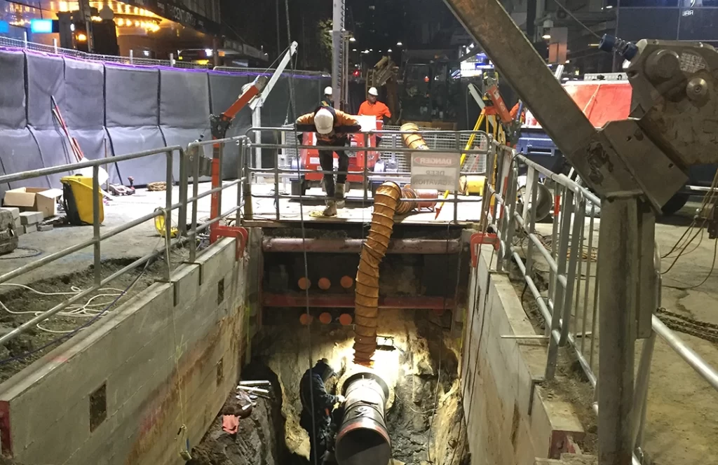 Two construction workers stand on a platform above a deep excavation site at night, supervising another worker inside the pit with large pipes and equipment.